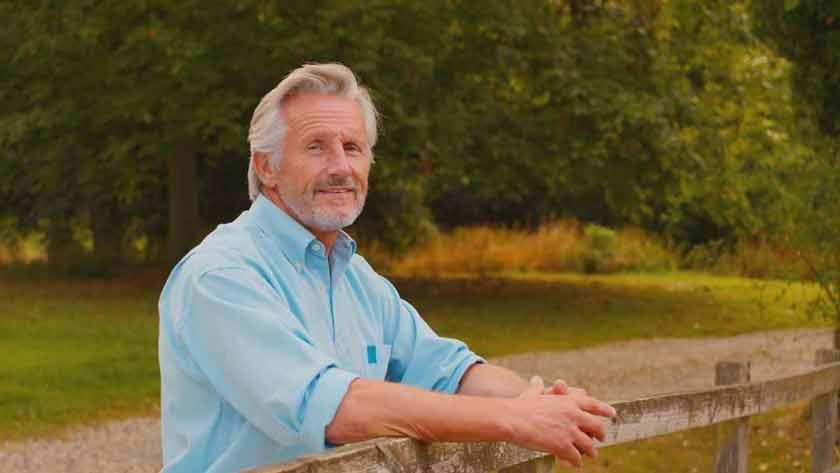 Portrait of an older man outdoors leaning on a wooden fence in a rural setting.